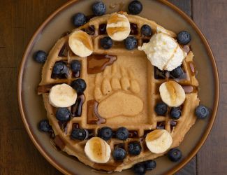 A waffle with "UM" baked in the middle is adorned with blueberries, butter and whipped cream