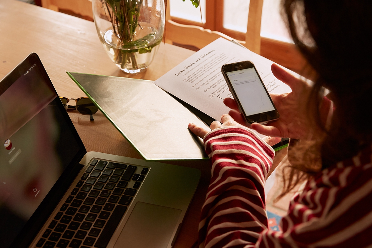 A person checking information on their laptop and their cell phone while referencing printed documents.