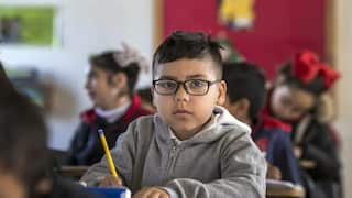 A boy in glasses sits at a school desk, holding a pencil. 