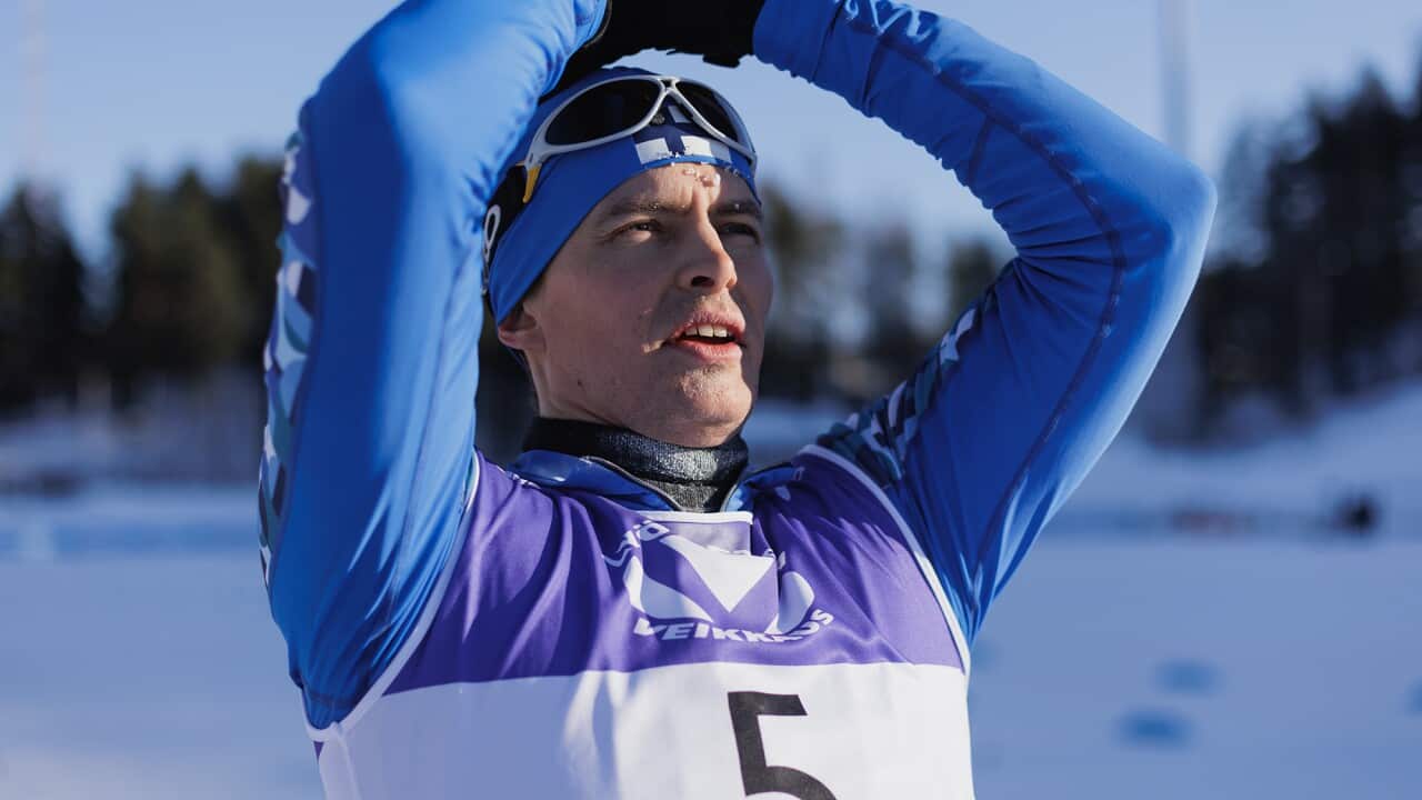 A man in a cross-country ski competitor's outfit stands in the snow, hands on his head. 