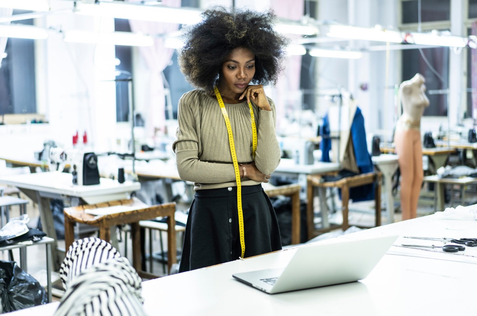 Image of a black woman fashion designer in a sewing room, looking at her computer screen with a pensive expression. She is working on product design strategy, reflecting consulting areas from strategic planning to product & marketing strategy.