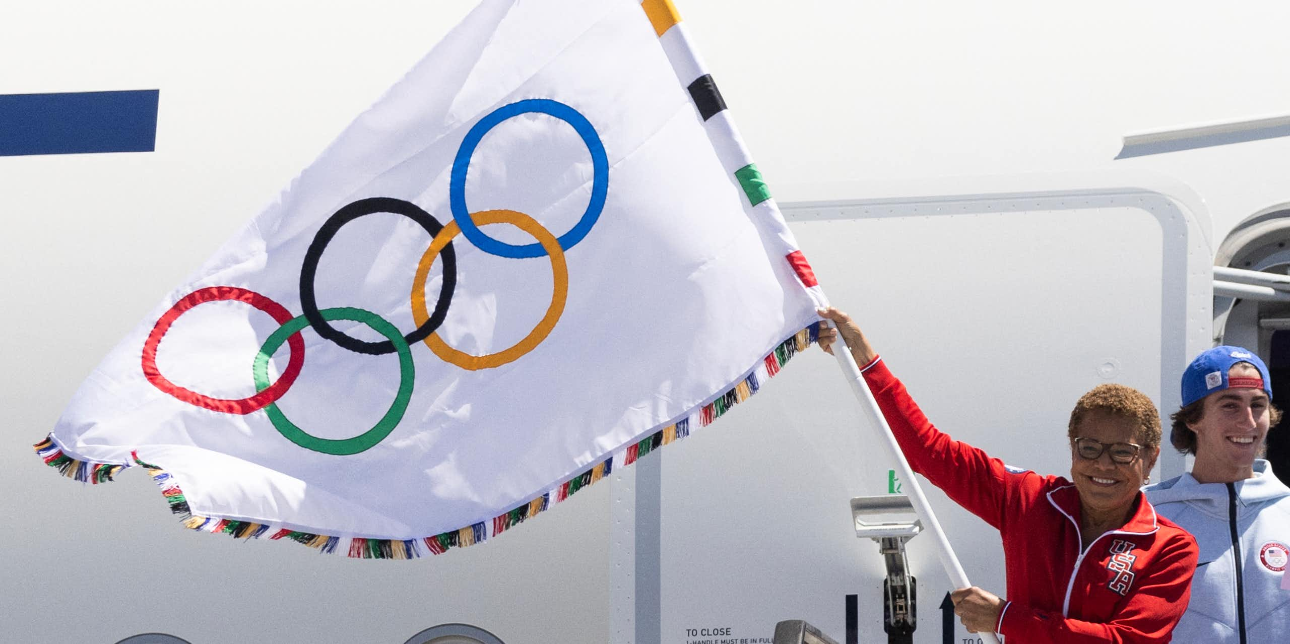 Une femme vêtue d'une veste d'entraînement rouge se tient à l'extérieur d'un avion à réaction en brandissant le drapeau olympique.