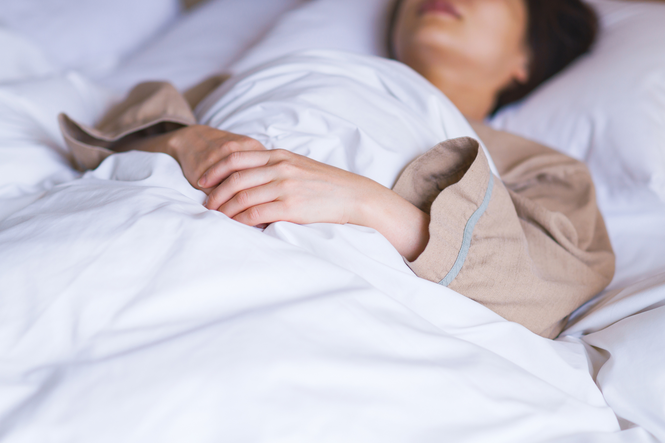 A person in a taupe robe lies under white bed sheets with hands folded on their stomach, face partially visible because of the angle