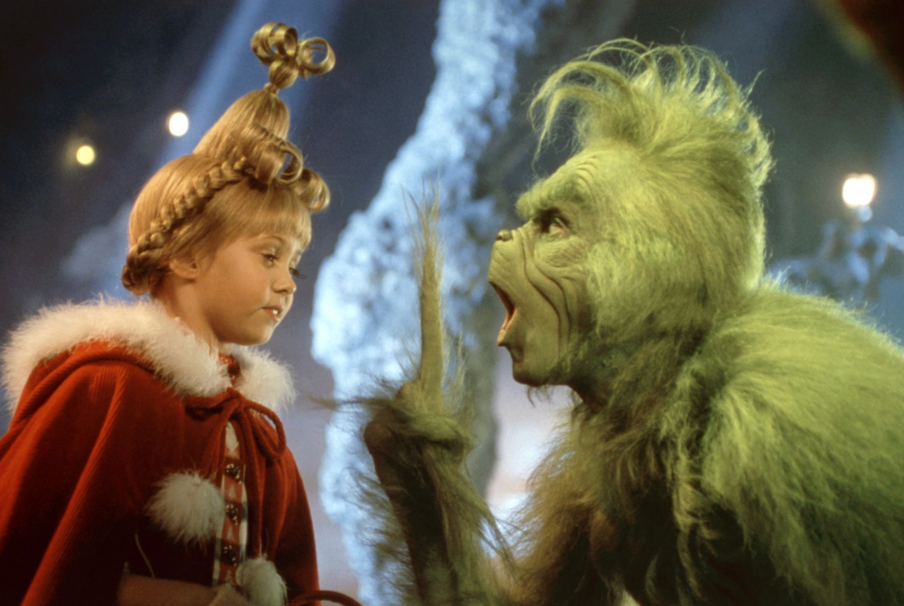 A child with an elaborate hairstyle looks at a furry, green creature. The child wears a cloak with a fur-trimmed hood