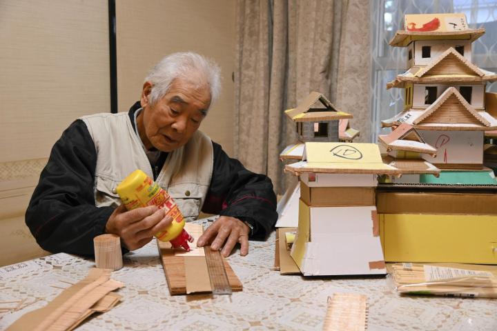 つまようじで松山城の模型（右）を制作する日野さん。ガムテープの粘着面に並べたつまようじに接着剤を塗り、薄い板状にしていく