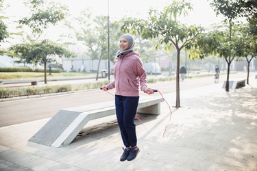 Person wearing a pink sweatshirt and black sweatpants jumping rope in a park outside.