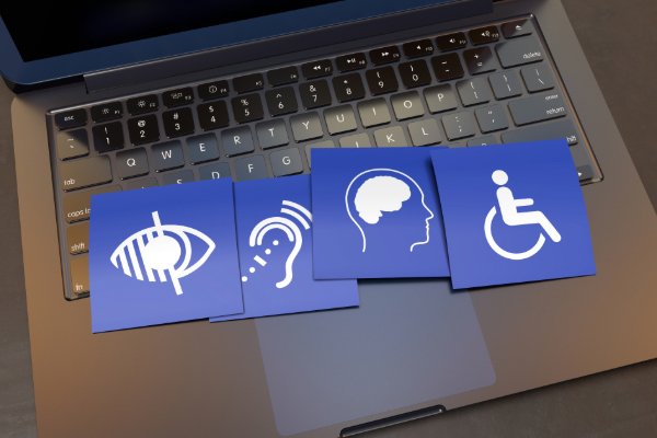 Four blue paper squares, each with a symbol or icon for a disability, lie on top of the keyboard of a laptop. The icons, from left, indicate sight, hearing, cognitive, and physical impairments.