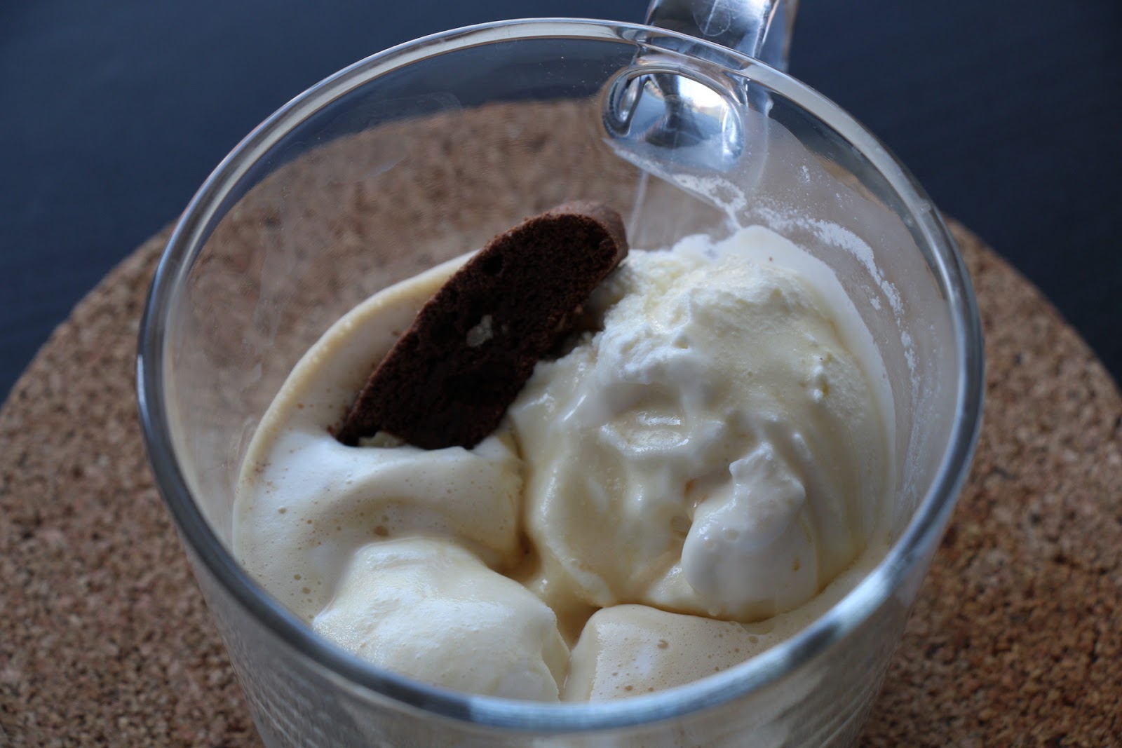 A dessert of affogato in a transparent glass cup, featuring creamy vanilla ice cream partially melted by a shot of hot espresso, topped with a chocolate biscotti.