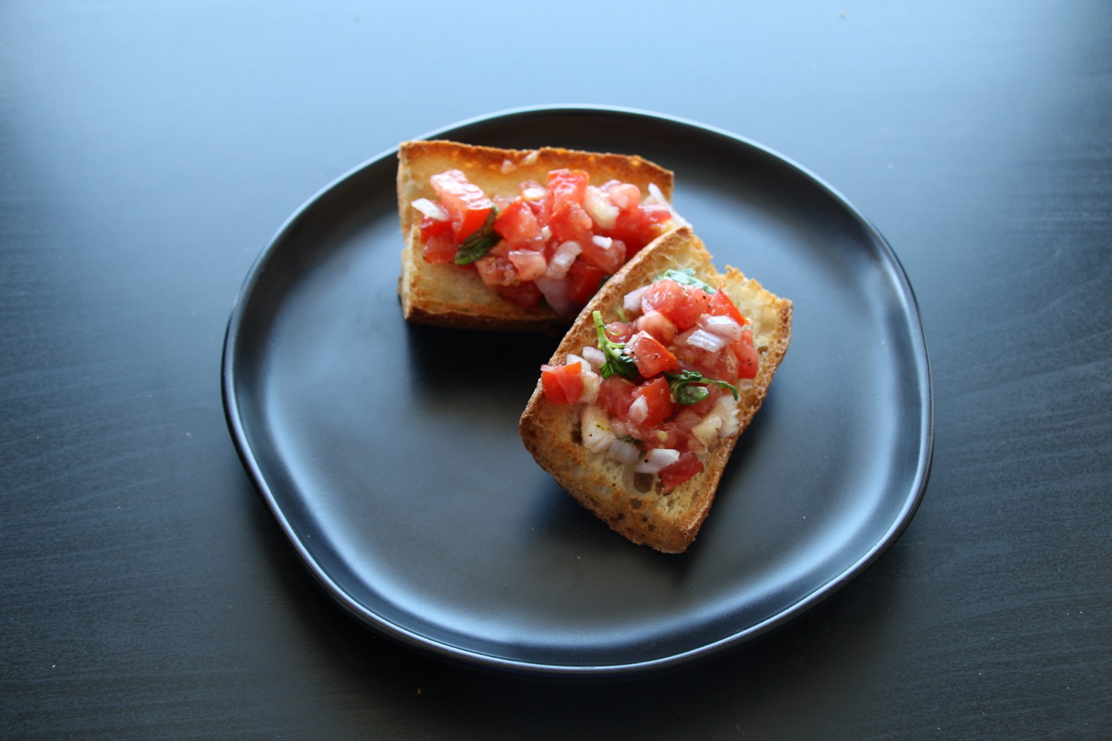 Two pieces of bruschetta on a black plate, each topped with diced tomatoes, onions, fresh basil, and a drizzle of olive oil on toasted bread slices.