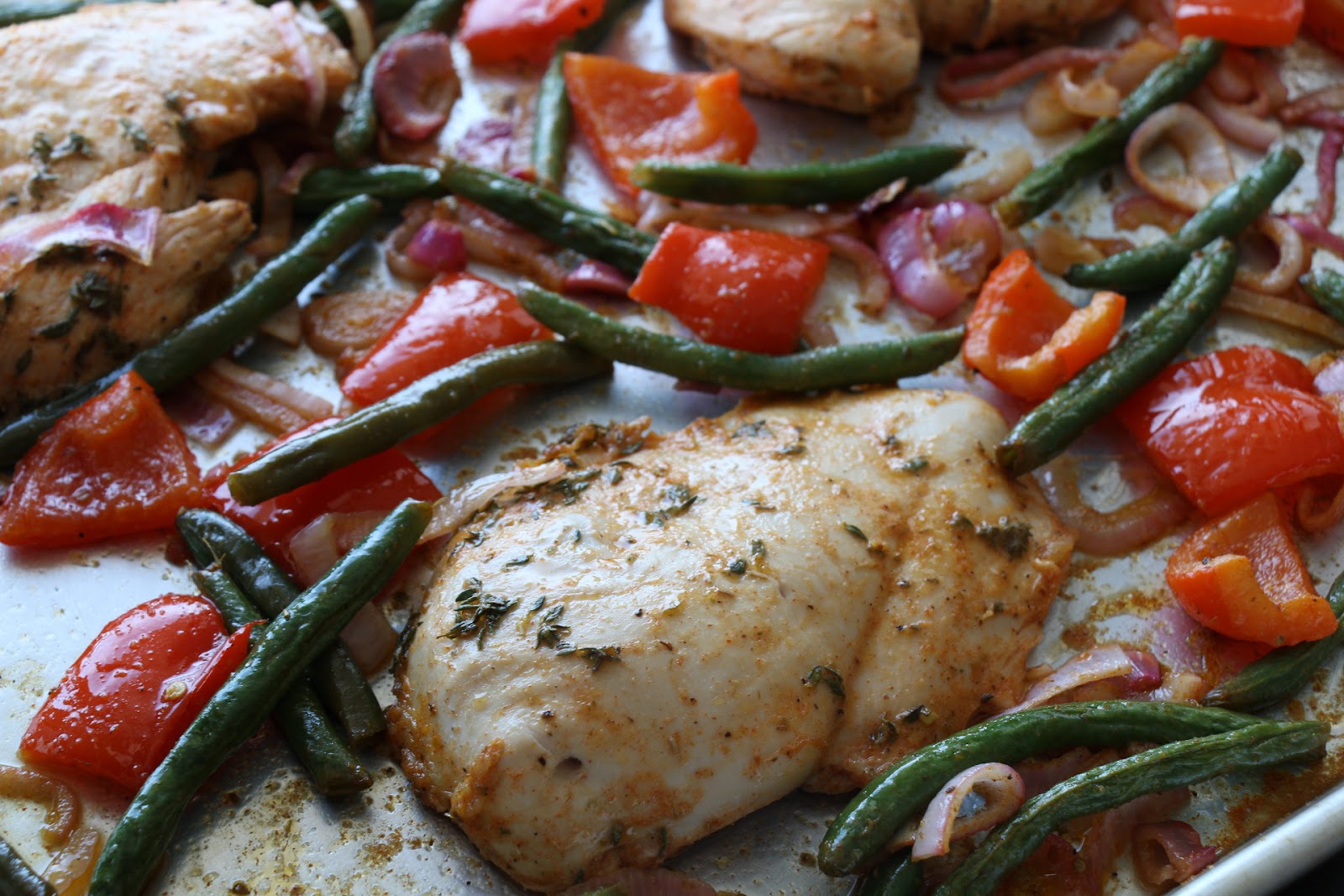 A sheet pan meal featuring seasoned chicken breasts, green beans, red bell pepper chunks, and sliced red onions, all roasted together with visible herbs.