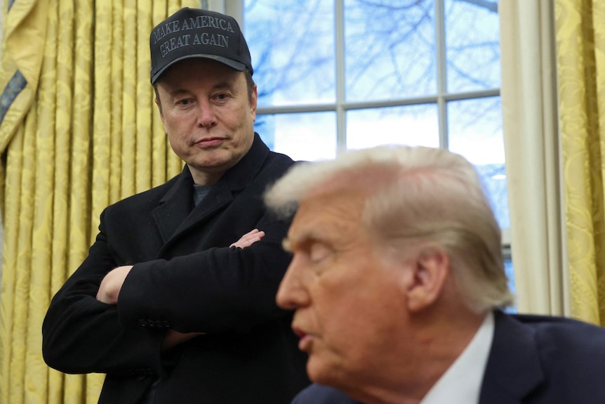 A man standing with his arms folded, looking at another man who is sitting and speaking