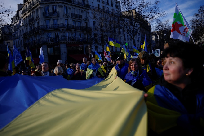 A crowd gathered around a huge yellow and blue Ukrainian flag in a street in front of an art deco building