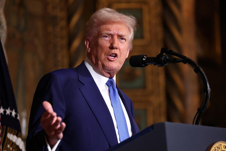 Donald Trump wearing a blue suit and tie gesturing with his right hand while standing at a US presidential lectern