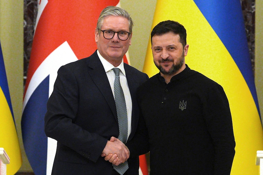 Keir Starmer and a smiling Volodymyr Zelenskyy shaking hands in front of UK and Ukrainian flags and two white lecterns