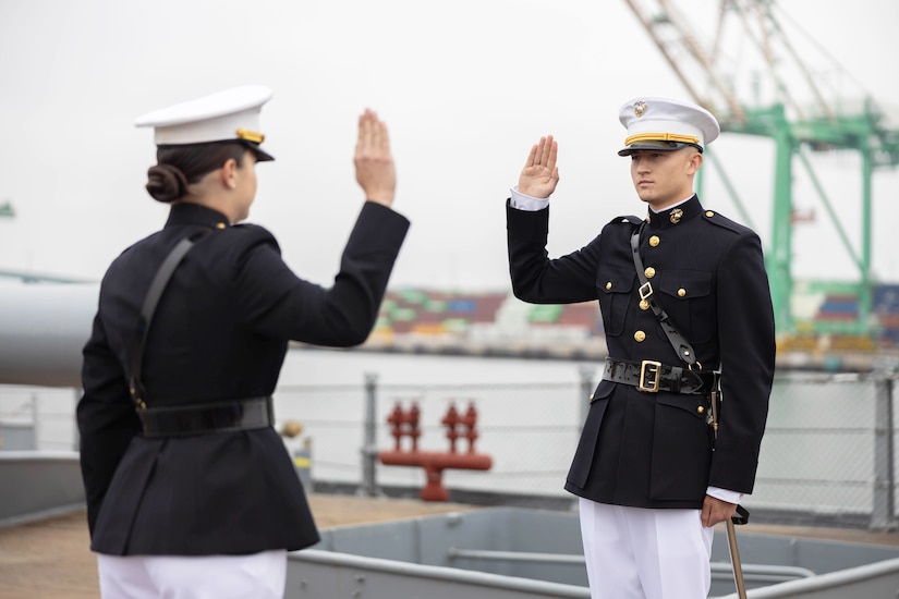 A Marine faces an officer as they both raise their right hands aboard a ship on a gloomy day.