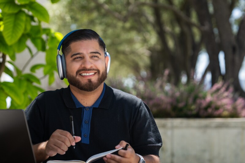 A person sits at a table with a computer and notebook, listening to something on their headphones 