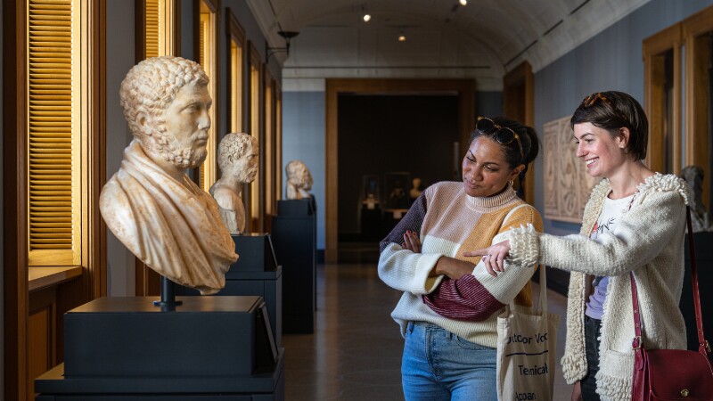 Two people wander through an art gallery filled with sculptures, pointing to the details in a stone bust