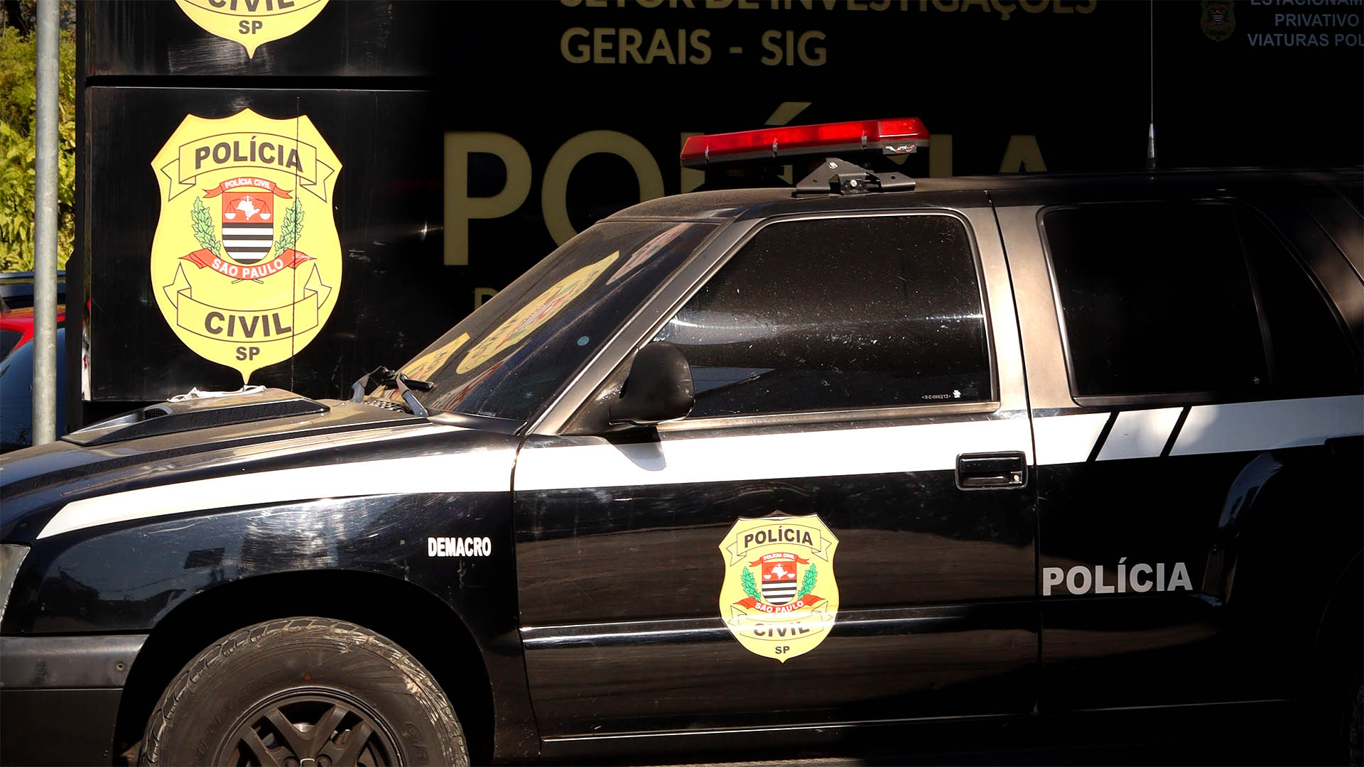 A black police car parked in front of a sign bearing the São Paulo police shield.