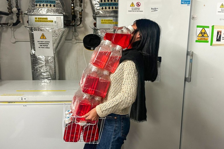 A lab worker carrying many bottles of red cell culture media past lab freezers and airducts
