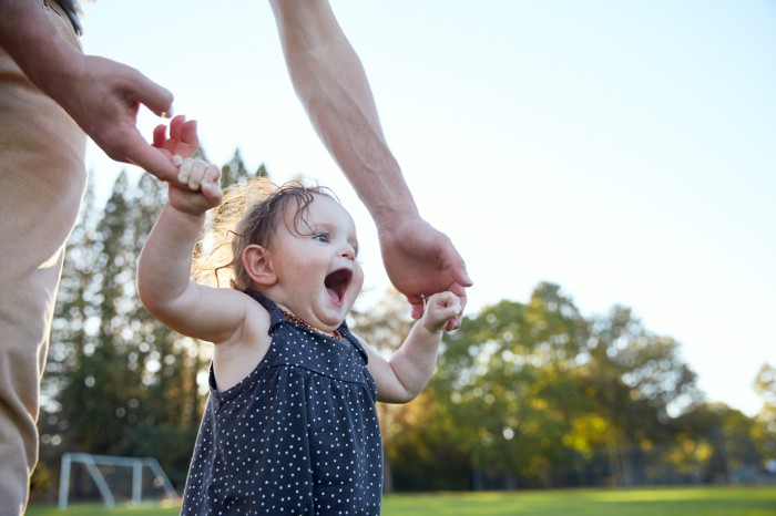 A baby with a big smile holds onto an adult's hands while learning how to walk.