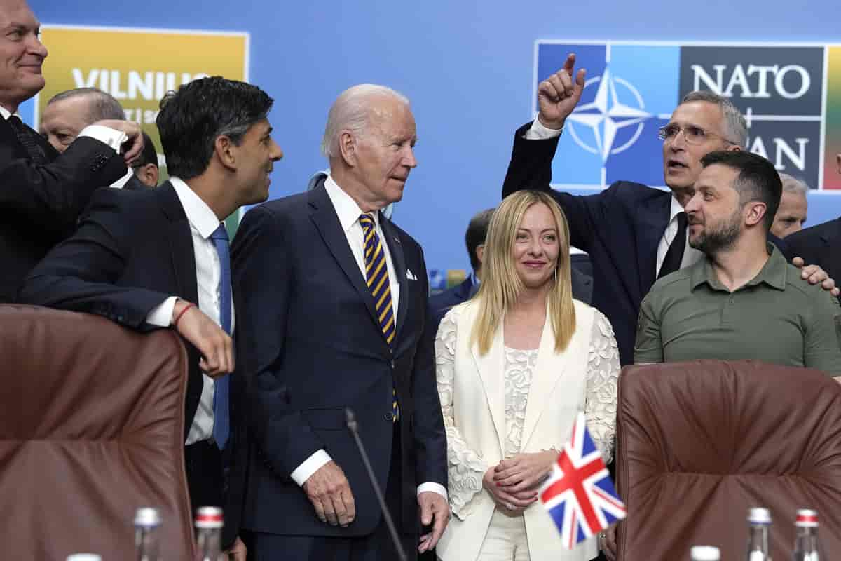 Fem menn og én kvinne inne i et konferanserom. Kvinnen, Meloni, har på en hvit dress med blomsterblonder. Zelenskyj har en stram militærgrønn genser med krage. De andre fire mennene har mørke dresser. Stoltenberg står med hånden på skulderen til Zelenskyj og peker opp i være med den andre. På vegge ibakgrunnen er NATOs logo. Foto