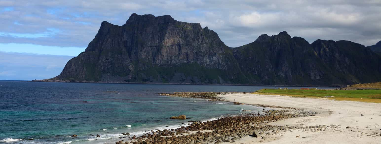 Foto med strand og hav i forgrunnen og bratte fjell i bakgrunnen