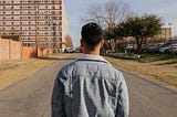 A man with short hair wearing a denim jacket. The man is facing away from the camera looking at an empty road ahead of him. There are buildings and a car park on the side of the road.