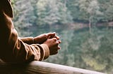 Man’s arm and hands, with man’s two hands together. Man is facing a background with a blurred image of bushes surrounding a body of water.