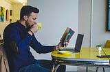 A man wearing a blue long-sleeved shirt, drinking from a yellow cup and reading a book. In front of the man is a laptop on a yellow dining table.