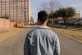 A man with short hair wearing a denim jacket. The man is facing away from the camera looking at an empty road ahead of him. There are buildings and a car park on the side of the road.