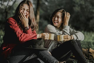 Two ladies sitting on the ground in a park, both holding a mug, both laughing