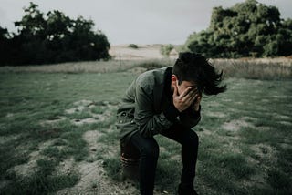 A young man wearing a green jacket and black pants seated in the middle of a park, both hands on the face almost covering it. There are 2 bushes in the background.