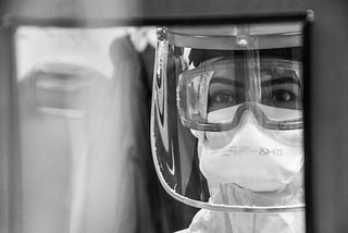 A healthcare worker in full protective gear, including a face shield, goggles, and a mask, looks through a window or screen with a focused and serious expression. The image is in black and white, highlighting the reflective surfaces and the clarity of the worker’s eyes.