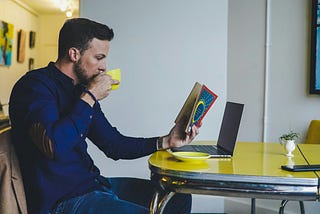 A man wearing a blue long-sleeved shirt, drinking from a yellow cup and reading a book. In front of the man is a laptop on a yellow dining table.