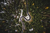 A helicopter view of a curved mountain road surrounded by green pine trees and a slight bit of snow.