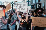 A group of people participating in a protest, holding signs that include messages such as ‘Protect Queer Youth’ and ‘Think of the Children! Not the Church.’ The protestors are wearing masks, and the atmosphere appears serious and determined as they advocate for LGBTQ+ youth rights.