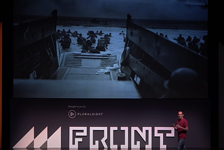 Wade Shearer on stage with a picture of soldiers running out of a boat towards the beach on D-Day in the background.