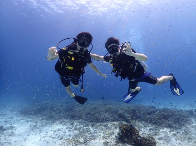 青い海に中で肩を組んだ男性2人がカメラに向かってハンドサインをしています。