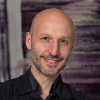 Glenn facing forward smiling, shaved head, black shirt, arms folded