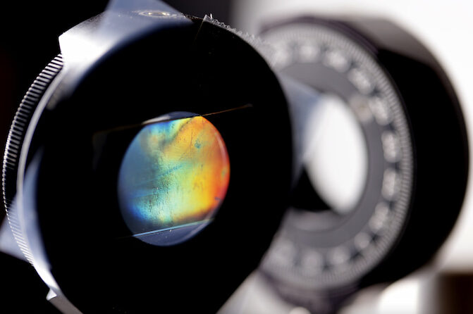 A thin sheet of material is sandwiched between two black, donut-shaped clamps. Through the hole, the sheet's shimmering color shifts from blue to red.