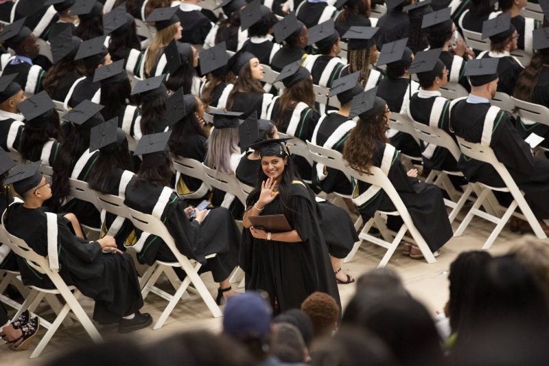 A UM graduate smiles and waves at the crowd during a convocation ceremony. Rows of grads in black caps and gowns face the stage behind her.