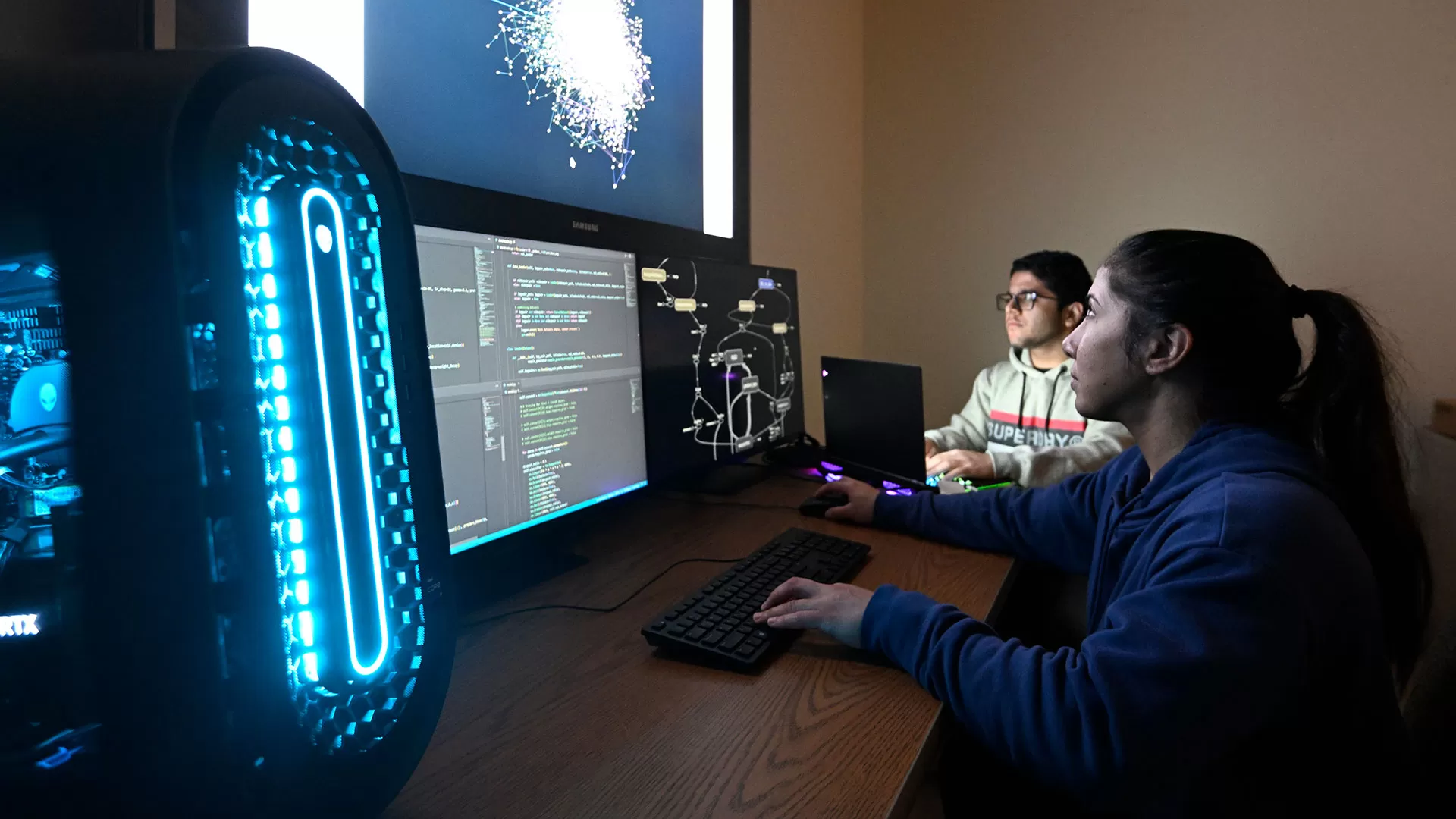 Two students work on computers while looking at a brightly lit screen.