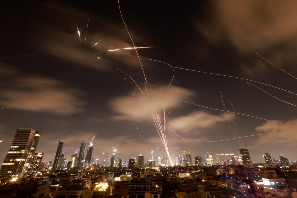 Israeli air defense systems intercept Iranian missiles over Tel Aviv at night, with multiple streaks of light visible in the cloudy sky above illuminated city buildings.