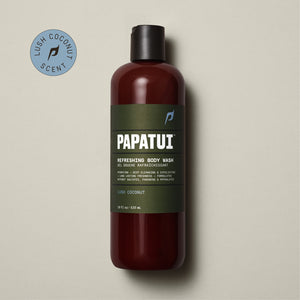 A PAPATUI Refreshing Body Wash in Lush Coconut scent in a translucent brown bottle with a black cap, displayed against a beige background with a few dark twigs. The label on the bottle is in a matching brown tone with white and orange text, highlighting features like hydrating, deep cleansing, exfoliating, and long-lasting freshness.