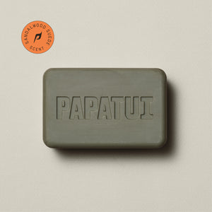 A bar Enriching Bar Soap in Cedar Sport scent rests on a beige background, with a sprig of greenery to the left. The soap is light beige and has a smooth texture with the PAPATUI brand name embossed on the surface. 