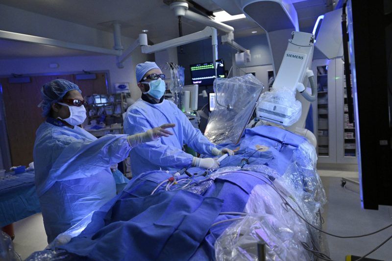 Two physicians perform a procedure on a draped patient in a surgical setting. Various pieces of medical equipment are located throughout the room.