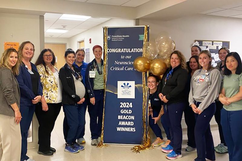 Thirteen members of the Neuroscience Critical Care team at Milton S. Hershey Medical Center smile as they stand around a pull-up banner that reads “Congratulations to our Neuroscience Critical Care – 2024 Gold Beacon Award Winners.”