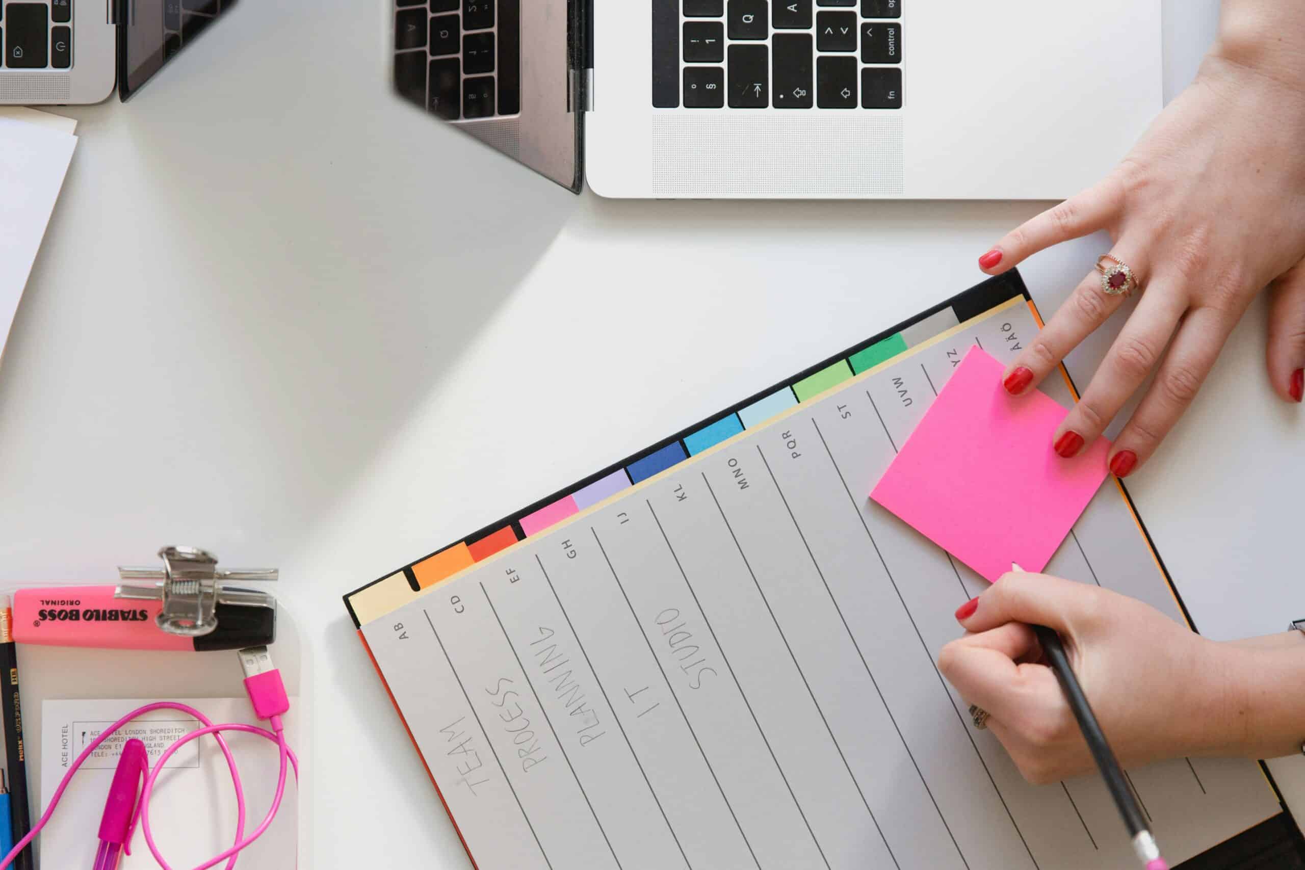 person writing a sticky note at a table with a planner and laptop