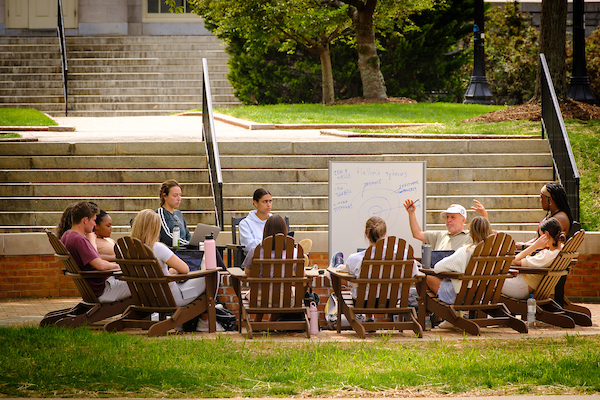 students sit in adirondack chairs around the professor outside on the Mag quad