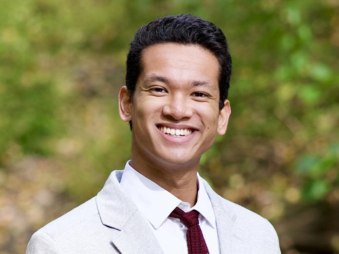 Young college-aged man with dark hair wearing a khaki suit. 